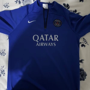 Blå PSG fotbollströja från Nike - Snygg blå Paris Saint-Germain fotbollströja från Nike med PSG-logga på bröstet och Qatar Airways tryck framtill. Tröjan har kort dragkedja vid halsen och är kortärmad. Tillverkad i lätt och funktionellt material som passar perfekt för träning eller match.