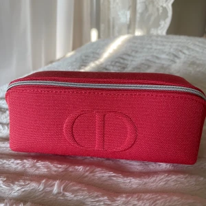 Röd necessär från D*or Beauté - Snygg röd necessär från C D  Beauté med broderad CD-logga på framsidan. Väskan är rektangulär och har en silverfärgad dragkedja runt om. Tillverkad i slitstark polyester med matchande röd insida. Perfekt för smink eller småprylar. Original, äkta. 