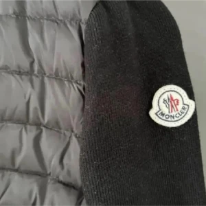 Svart pufferjacka från Moncler - Svart pufferjacka från Moncler med quiltad front och stickade ärmar. Jackan har dragkedja framtill, två fickor och klassisk Moncler-logga på ärmen. Perfekt för dig som vill ha en stilren och sportig look.