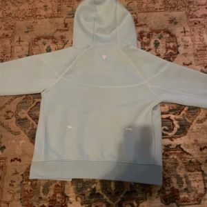 Nike NOCTA ljusblå hoodie med dragkedja - Säljer en sjukt snygg ljusblå hoodie från Nike NOCTA med hel dragkedja och huva. Tröjan har två fickor med dragkedja framtill, diskreta vita loggor och coola detaljer på ärmen. Perfekt för dig som gillar streetwear och vill sticka ut lite extra.