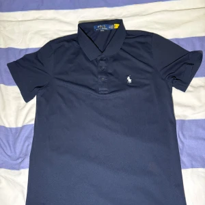 Mörkblå piké från Polo Ralph Lauren - Snygg mörkblå pikétröja från Polo Ralph Lauren med klassisk krage och tre knappar framtill. Ikoniska broderade loggan i vitt på bröstet. Tröjan är kortärmad och gjord i mjuk bomull som är skön mot huden. Perfekt för dig som gillar stilrena och tidlösa plagg.