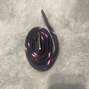 Svart flätat bälte med röd, blå och vit detalj - Snyggt svart flätat bälte med inslag av röda, blå och vita ränder. Bältet har en metallspänne och detaljer i mörkbrunt läder vid ändarna. Perfekt accessoar för att lyfta din outfit med lite färg och stil.