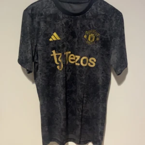 Manchester United svart tränings-tshirt - Snygg svart Manchester United tränings-tshirt från Adidas med guldiga detaljer och logga. Tröjan har korta ärmar och ett marmorerat mönster över hela tyget. Materialet är lätt och andas, perfekt för fotbollsträning eller häng med kompisar.