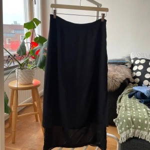 Svart långkjol med slits och dragkedja - Svart långkjol i flowigt, lätt material med transparent nederdel och snygg slits på sidan. Kjolen har en diskret dragkedja i midjan för enkel på- och avtagning. Perfekt för dig som gillar minimalistisk och stilren look.