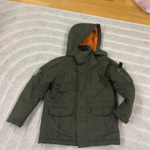 Grön parkajacka från KappAhl  - Parkajacka till barn. I mörkgrönt med orange foder,  huva och flera fickor. Jackan har patchar med text på ärmen och ryggen, dragkedja och knäppning framtill samt slits bak. Perfekt för kalla dagar och riktigt snygg streetstil.