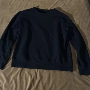 Mörkblå sweatshirt från Polo Ralph Lauren - Mörkblå sweatshirt från Polo Ralph Lauren i storlek XL. Tröjan har en klassisk rund hals och långärmade ärmar. På bröstet finns den ikoniska röda Polo-loggan broderad. Materialet är mjuk bomull som känns skön mot huden.