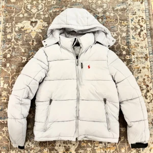 Vit pufferjacka från Polo Ralph Lauren - Säljer en fräsch vit pufferjacka från Polo Ralph Lauren med huva och dragkedja. Jackan har två sidofickor med dragkedja och en liten röd Polo-logga på bröstet. Perfekt för kalla dagar och har en modern, clean look.
