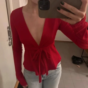 Röd omlottopp med knyt och v-ringning - Säljer en snygg röd omlottopp med djup v-ringning och långa ärmar. Toppen har ett ribbat tyg och knytband i midjan som ger en schysst siluett. Perfekt för dig som gillar färg och vill sticka ut lite extra.