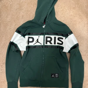 Grön Paris Saint-Germain Jordan hoodie - Snygg grön hoodie från Jordan x Paris Saint-Germain med stor PARIS-logga och Jumpman-tryck över bröstet. Huva, dragkedja och kängurufickor framtill. Vita och svarta detaljer på ärmarna samt PSG-märke på ena ärmen. Tillverkad i mjuk bomullsmix.