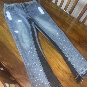 Gallery dept - Snygga grå jeans med vita och svarta färgstänk längs benen och slitna detaljer upptill. Byxorna har en rak passform och råa kanter vid bensluten. Materialet är klassisk denim och färgen är ljusgrå med inslag av svart längs sidorna.