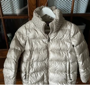 Beige dunjacka från Colmar, 130 cm barnstorlek - Snygg beige dunjacka från Colmar med quiltad design och hög krage. Jackan har dragkedja framtill och två fickor med dragkedja. Perfekt för kyliga dagar och ger en clean look med sin glansiga finish. Liten i storlek 