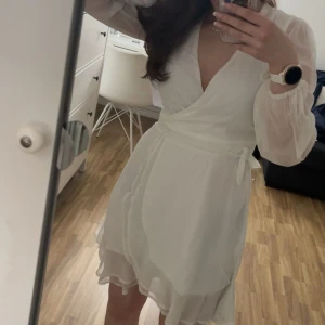 Vit omlottklänning från Bubbleroom - Supersöt vit omlottklänning från Bubbleroom med långa transparenta ärmar och v-ringad hals. Klänningen har volangdetaljer nertill och ett knytband i midjan som ger en snygg siluett. Helt oanvänd med prislapp kvar. Öppen för prisförslag.
