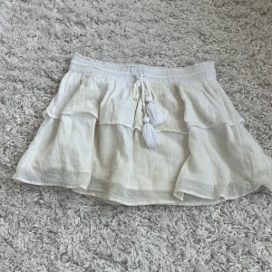 Vit volangkjol från Gina Tricot - Söt vit kortkjol från Gina Tricot med volanger och knytband med tofsar i midjan. Kjolen har en luftig och flowy känsla, perfekt för varma dagar. Elastisk midja för skön passform och tillverkad i mjukt bomullstyg.