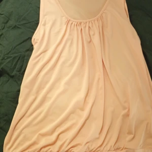 Ljus persikofärgad ärmlös klänning 1XL - Säljer en ljus persikofärgad ärmlös klänning i storlek 1XL. Klänningen har rundad halsringning, lätt rynkade detaljer framtill och en lös, flowy passform. Perfekt för varma dagar och enkel att styla med accessoarer.
