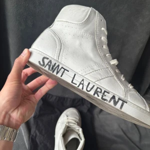 Saint Laurent vita high-top sneakers - Snygga vita high-top sneakers från Saint Laurent med handskriven logga på sulan. Skorna är tillverkade i mjukt skinn och har klassisk snörning samt rund tå. YSL-mönster på undersidan av sulan ger extra lyxig känsla. Perfekt för dig som gillar stilrena och exklusiva sneakers.