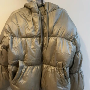 Beige pufferjacka från H&M Divided - Säljer en beige pufferjacka från H&M Divided med huva och dragkedja framtill. Jackan har en oversized look, två stora fickor och snörning vid huvan. Materialet är syntet och jackan är perfekt för kalla dagar.