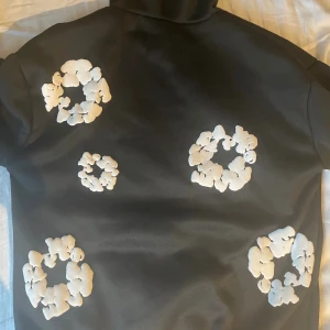 Denim tears hoodie - Cool svart hoodie med unika vita blommönster över hela tröjan, även på huvan och ärmarna. Tröjan har en klassisk huva och magficka framtill. Materialet känns mjukt och har en lite tjockare känsla, perfekt för att hålla värmen. Passformen är normal och designen sticker verkligen ut. Svart hoodie med vita blommönster