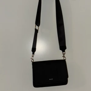 Svart axelväska från NA-KD - Stilren svart axelväska från NA-KD med bred justerbar axelrem och silverfärgade metalldetaljer. Väskan har ett lock med magnetstängning och ett huvudfack med dragkedja. Perfekt storlek för det viktigaste och enkel att matcha till olika outfits.