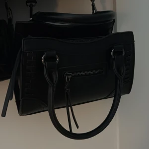 Svart handväska från Gina  - Snygg svart handväska i skinn med stilrena detaljer och silverfärgade metalldelar. Väskan har rundade handtag och en praktisk dragkedja framtill. Designen är klassisk med diskreta mönster längs sidorna och en strukturerad form som ger en cool vibe.
