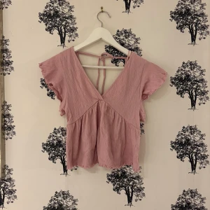 Rosa blus med volangärm från lager 157 - Supersöt ljusrosa blus från lager 157 med v-ringning och knyt i ryggen. Blusen har korta volangärmar och är tillverkad i ett mjukt, krinklat bomullstyg som ger en chill och feminin vibe. Perfekt till jeans eller kjol för en söt look.