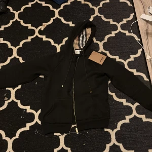 Svart Burberry hoodie med dragkedja - Svart hoodie från Burberry med klassiskt rutigt foder i huvan och guldfärgade detaljer på snörningen. Tröjan har dragkedja framtill, fickor och en snygg, stilren look. Perfekt för dig som vill ha något exklusivt och bekvämt.