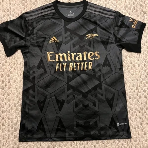 Arsenal svart bortatröja Adidas - Snygg svart Arsenal bortatröja från Adidas med guldiga detaljer och geometriskt mönster. Tröjan har korta ärmar, rund hals och är tillverkad i lätt och ventilerande AEROREADY-material. Perfekt för dig som vill sticka ut på läktaren eller planen.
