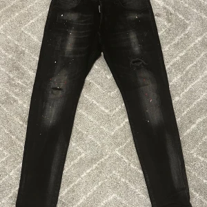 Svarta Dsquared2 jeans med färgstänk - Snygga svarta jeans från Dsquared2 med slitningar och coola färgstänk i rött och vitt. Jeansen har en smal passform och klassisk femficksdesign. Märkespatch bak i midjan med Dsquared2-logga och lönnlöv. Perfekta för dig som vill sticka ut med din stil.