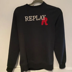 Svart sweatshirt från Replay med tryck - Svart sweatshirt från Replay i storlek M med REPLAY-tryck i vitt och en röd broderad R på bröstet. Tröjan har rund hals, långa ärmar och ribbade muddar. Materialet känns som mjuk bomull och passformen är normal. Perfekt för dig som gillar stilrena logotröjor. Väldigt bra skick förutom hålet i armhålan men den märks inte 