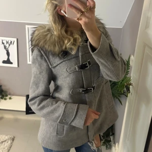 Grå kappa med päls från Zara Woman - Snygg grå kappa från Zara Woman med fluffig päls runt huvan och coola spänndetaljer framtill. Jackan har lång ärm, huva och en loose passform. Perfekt för dig som vill ha en stilren och trendig look under höst och vinter.