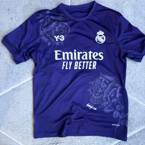 Real Madrid  Y-3  - Säljer en lila Real Madrid Y-3 fotbollströja från Adidas med vita tryck och ros-mönster. Tröjan har korta ärmar. Skriv för mer information eller on priset. Storleken är M.