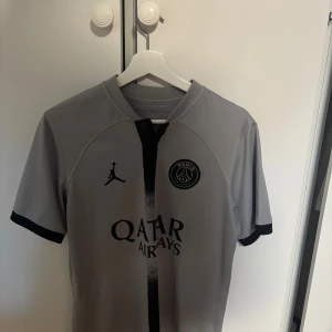 PSG grå fotbollströja  - Snygg grå PSG fotbollströja med svart vertikal rand och svarta detaljer. Tröjan har korta ärmar, Paris Saint-Germain-logga på bröstet och Jordan-logga. Tillverkad i lätt och ventilerande material, perfekt för match eller träning.
