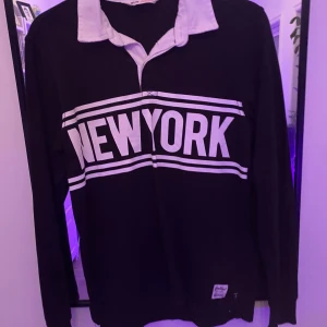 Svart pikétröja med New York-tryck H&M - Svart långärmad pikétröja från H&M med vit krage och stort New York-tryck över bröstet. Tröjan har vita ränder och klassisk pikédesign med knappslå. Perfekt för dig som gillar sportig och urban stil.