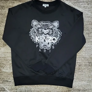 Svart sweatshirt från Kenzo med tiger - Svart sweatshirt från Kenzo i storlek M med ikonisk broderad tiger och Kenzo Paris-text framtill. Tröjan har rund hals, ribbade muddar och lång ärm. Materialet är mjukt och känns som bomull och polyester. Perfekt för dig som gillar statement-plagg och snygga detaljer.