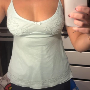 Ljusgrön linnetopp med pärlor från Lindex - Supersöt ljusgrön linnetopp från Lindex i storlek XS. Toppen har tunna axelband, broderade pärlor och hjärtan på bröstet samt en liten rosett i mitten. Materialet är mjukt och lätt, perfekt för varma dagar. Nedre kanten har dekorativa pärlor för extra detalj.