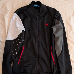 Svart Adidaströja med röda detaljer - Adidaströja i svart med grå och vita paneler på ärmarna, röda dragkedjor och röd Adidas-logga på bröstet. Ena ärmen har vita symboler som ger en unik touch. Jackan har ståkrage och hel dragkedja framtill. Katt finns i hemmet.