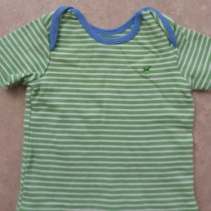 T-shirt - Snygg grön och vit randig t-shirt med blå kant runt halsen. T-shirten har korta ärmar och ett litet broderat motiv på bröstet. Perfekt för dig som gillar färgglada och lekfulla plagg.