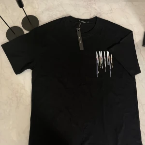 Svart AMIRI t-shirt med tryck - Svart t-shirt från AMIRI i bomull med coolt tryck framtill och baktill. Trycket består av AMIRI-loggan med färgstänk och droppande effekt i vitt, lila, gult och rött. Klassisk rund hals och korta ärmar. Perfekt för dig som gillar streetwear och statement-plagg.