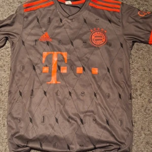 Bayern München Musiala fotbollströja - Säljer en grå kortärmad Bayern München fotbollströja från Adidas med Musiala och nummer 42 på ryggen. Tröjan har orange detaljer, ränder på axlarna och ett subtilt rutmönster med små symboler. Materialet är lätt och andas, perfekt för match eller träning.