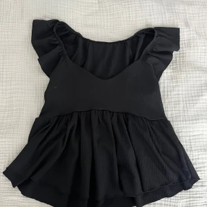 Svart blus med volangärmar - Säljer en svart blus med breda volangärmar och v-ringad hals. Toppen har en luftig, rynkad nederdel som ger en flowy känsla. Materialet är mjukt och lätt, perfekt för dig som gillar en feminin och chill stil. Passar till jeans eller kjol för en snygg look.