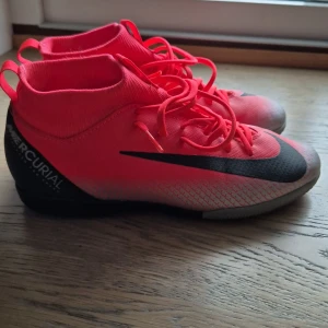 Nike Mercurial fotbollsskor röd/svart - Säljer ett par Nike Mercurial fotbollsskor i en riktigt snygg röd färg med svarta detaljer och logga. Skorna har snörning, platt sula och en strumpliknande högre krage för extra stöd. Materialet är syntet och designen är modern med mönstrad ovandel.