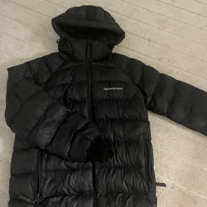 Svart Peak Performance dunjacka - Säljer en svart dunjacka från Peak Performance med huva och hel dragkedja. Jackan har en quiltad design, två sidofickor med dragkedja och lång ärm med muddar. Perfekt för kalla dagar och har en clean, sportig look.