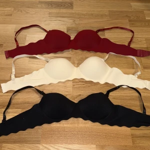 3-pack bh i olika färger - Aldrig använt för små för mig, Säljer ett 3-pack bh i vinröd, vit och svart. Alla har avtagbara axelband, vadderade kupor och vågig kant nedtill. Tillverkade i ett stretchigt syntetmaterial som andas. Perfekt för att bära under kläder eller som en enkel topp till sommaren.
