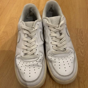 Nike Air Force 1 vita sneakers - Klassiska Nike Air Force 1 sneakers i helvitt skinn med vit sula och snörning. Ikonisk design med perforerad tå och Nike-logga på sidan och hälen. Perfekta för dig som gillar streetstyle och tidlös look.