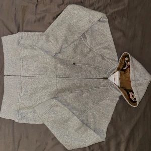 Grå hoodie från Burberry med dragkedja - Säljer en grå hoodie från Burberry med klassiskt rutigt foder i huvan och broderad logga på bröstet. Tröjan har hel dragkedja, snörning i huvan och är tillverkad i mjuk bomullsmix. Priset inte hugget i sten