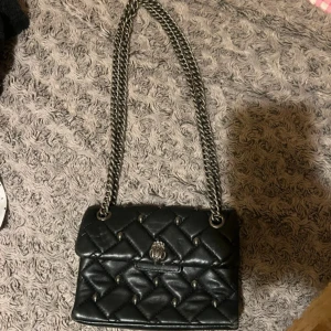 Svart quiltad axelväska Kurt Geiger - Snygg svart axelväska från Kurt Geiger i quiltat skinn med silverfärgade kedjor och detaljer. Väskan har ett coolt mönster med små nitar och en ikonisk logga framtill. Perfekt storlek för det viktigaste och ger en edgy vibe till din outfit.