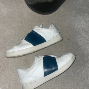 Valentino Garavani Open sneakers vit/blå - Snygga Valentino Garavani Open sneakers i vitt skinn med bred blå läderdetalj över vristen. Klassisk rund tå, platt sula och coola nitar längs hälen. Perfekta för dig som vill ha en lyxig och stilren look med en twist. Dom är nya utan prislapp använda 3 - 5 gånger bara. Storlek 44 men passar 43 bra.