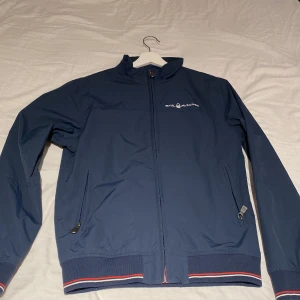 Marinblå vindjacka Sail Racing S - Snygg marinblå vindjacka från Sail Racing med dragkedja framtill och två fickor med dragkedja. Jackan har ribbade muddar med röd och vit rand, hög krage och diskret logga på bröstet samt ryggen. Perfekt för blåsiga dagar och sportig stil. Strlk s 