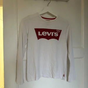 Vit långärmad Levi's tröja med tryck - Säljer en vit långärmad tröja från Levi's med det klassiska röda logotrycket på bröstet. Rund hals med röd detalj på insidan av kragen. Perfekt för dig som gillar streetwear och ikoniska märken.