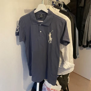 Blå pikétröja från Polo Ralph Lauren - Snygg blå pikétröja från Polo Ralph Lauren med klassisk krage och två knappar framtill. Stor vit broderad polospelare på bröstet och en stor vit siffra på ärmen. Tröjan är gjord i mjuk bomull och har korta ärmar, perfekt för en chill och stilren look. Storlek S. Tajt runt armarna!