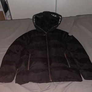 Svart pufferjacka från Moncler - Säljer en svart pufferjacka från Moncler med stor huva och tydlig logga på ärmen. Jackan har dragkedja framtill och två sidofickor med dragkedja. Moncler-tryck på huvan ger en extra cool vibe. Perfekt för kalla dagar och snygg streetstil.